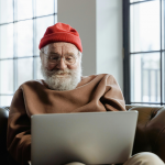 Image of elderly man with red hat, sitting on a couch and looking at an open laptop.